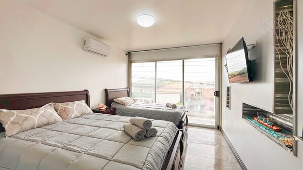 A bedroom with two beds, a TV mounted on the wall, and a sliding glass door leading to a balcony.