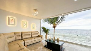 Una sala de estar moderna con un sofá de cuero color beige, una mesa de centro de vidrio y obras de arte enmarcadas en la pared. Se ve una palmera a través del amplio ventanal con vistas al mar.