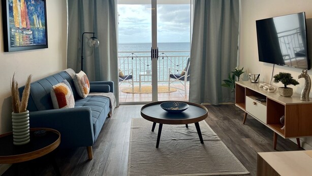 Ein Wohnzimmer mit einem blauen Sofa, einem runden Couchtisch, einem Fernseher und einem Balkon mit Meerblick.