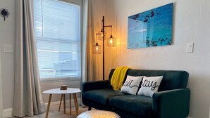Una sala de estar acogedora con un sofá verde oscuro, dos cojines blancos con el texto 'sit stay', un plaid amarillo, un reposapiés plateado redondo, una mesita blanca pequeña y una lámpara de pie.