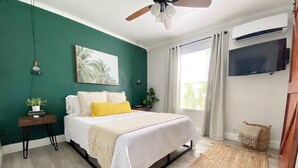 Un dormitorio con cama, ventilador de techo, televisor montado en la pared, ventana con cortinas y suelo de madera.