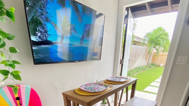 Un comedor con una mesa y sillas de madera, una silla de playa colorida, un televisor montado en la pared y vistas al patio trasero a través de una puerta abierta.