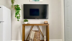 Una sala de estar con un televisor montado, una mesa consola de madera con una cesta tejida, una planta en maceta y un letrero enmarcado.