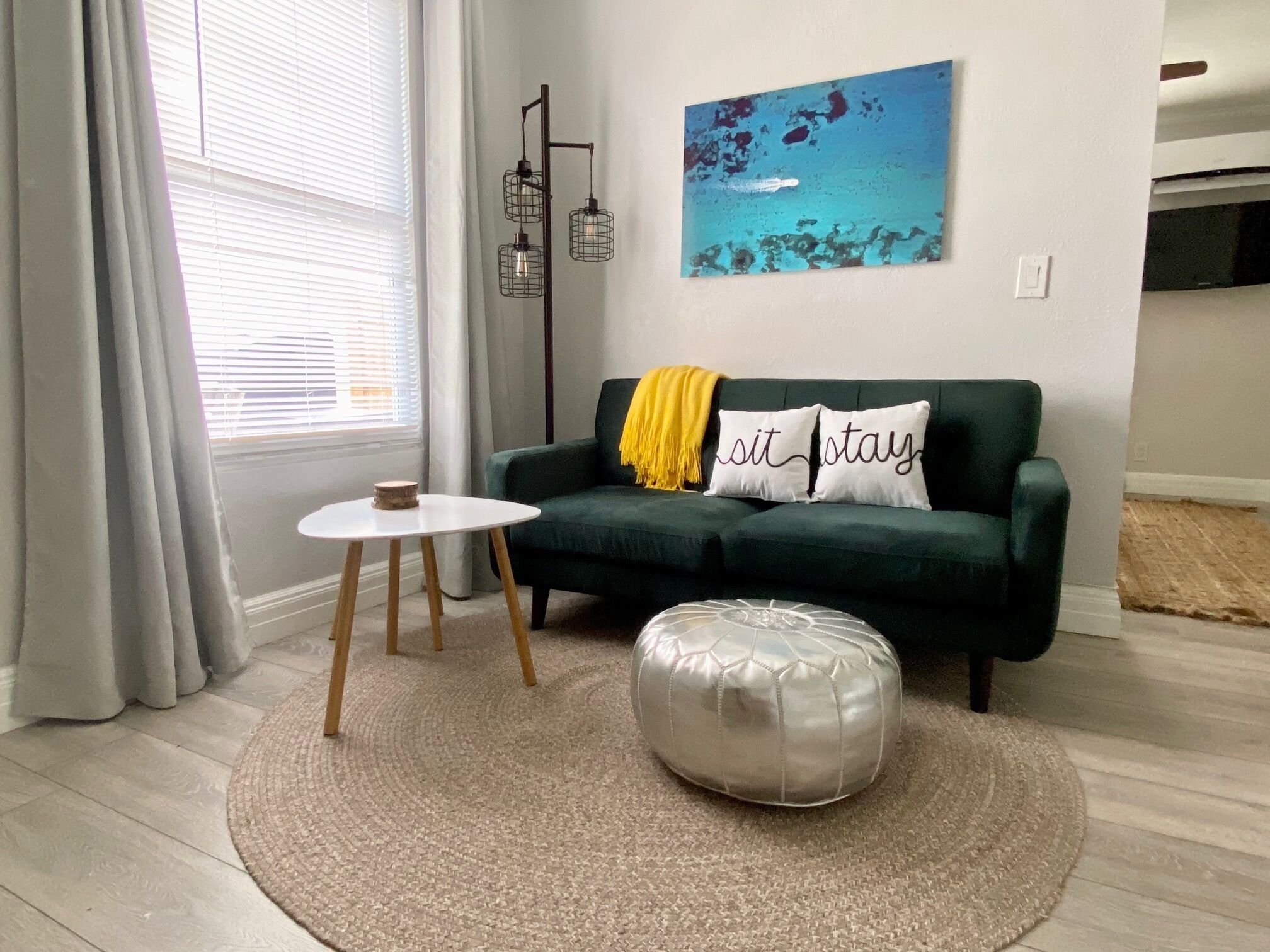 A modern living room with a green sofa, two white cushions, a round ottoman, a small white table, and a floor lamp.