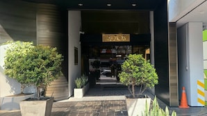 L’entrée d’un hôtel, avec une enseigne indiquant « AKROS ». De part et d’autre de l’entrée, on trouve des plantes en pot, un chemin pavé menant à l’intérieur et, sur la droite, un mur végétal orné de motifs géométriques.