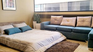 Un salon moderne avec un lit, un canapé gris et un petit fauteuil. Une grande fenêtre est agrémentée de rideaux légers, et on y trouve également une plante en pot et un tableau encadré accroché au mur.