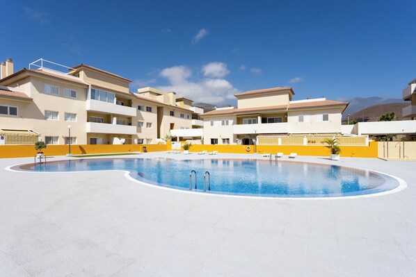 A swimming pool surrounded by multi-story buildings with balconies, a clear sky, and a mountainous background.
