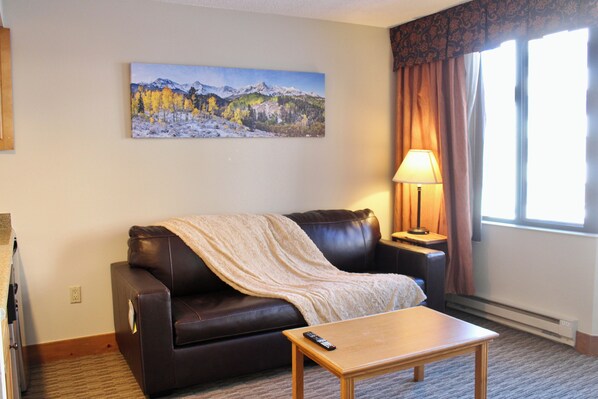 A living room with a brown leather sofa, a wooden coffee table, a remote control, a lamp, and a painting of a mountain landscape. - Walk to Mountain Base Studio for 4 & HotTub (Crested Butte)