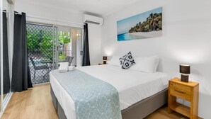 A bedroom with a bed, a nightstand, a wall-mounted air conditioner, a window with curtains, and a painting of a beach scene.
