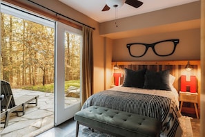 A bedroom with a large bed, a view of a forest through a sliding glass door, a ceiling fan, and a nightstand with a red lamp.