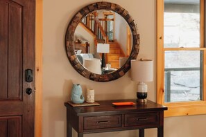 A wooden door, a round mirror with a rustic frame, a small table with a lamp, and a window with a view of a snowy landscape. - Secluded Catskills Treehouse | Hot Tub + Roof Deck (Pond Eddy)