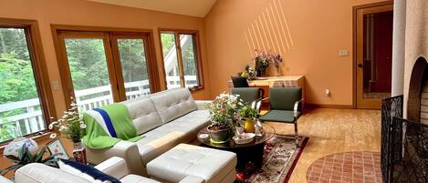 A spacious living room with a large sofa, a coffee table, and a fireplace. There are skylights on the ceiling, and the room has large windows.