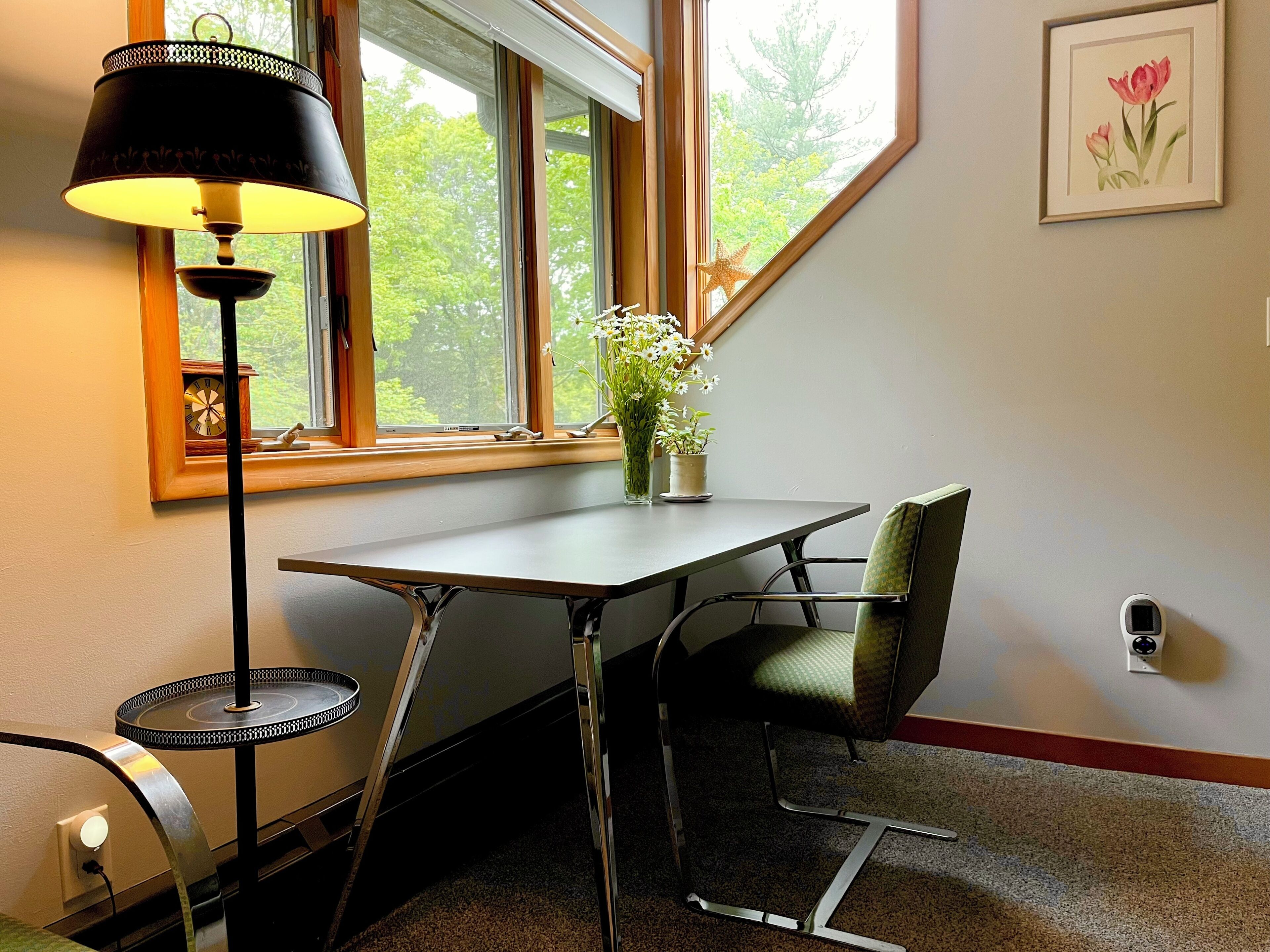 Une pièce d’angle avec une table, une chaise, une lampe, une fenêtre et un tableau encadré au mur.