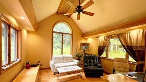 A spacious room with a high ceiling, a ceiling fan, a large window with a view of greenery, a white sofa, a black leather chair, and a wooden table with a computer setup. - Sunny Peace Homestay (Harwinton)
