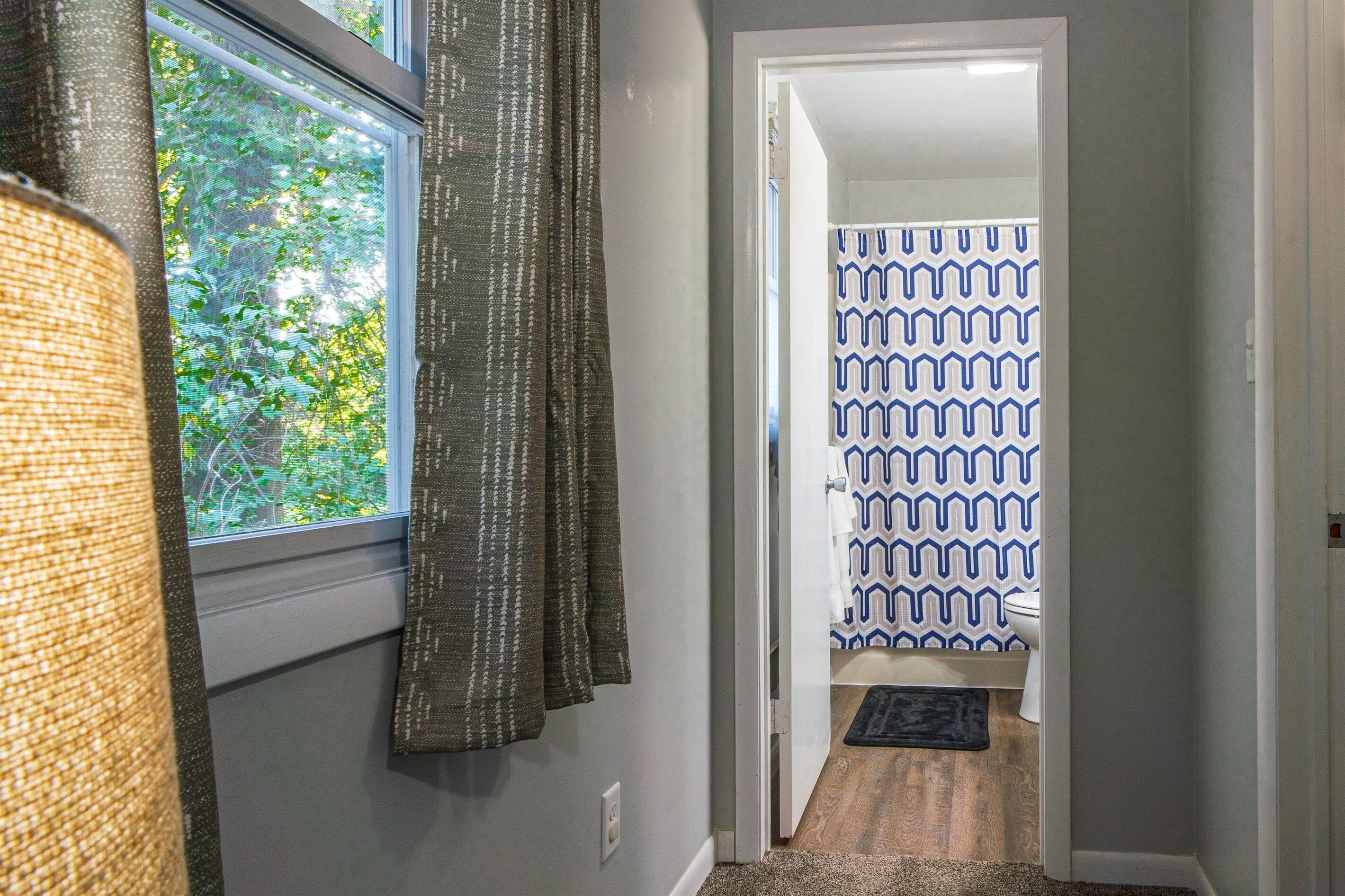 A hallway with a window, a door leading to a bathroom with patterned shower curtains, and a rug on the floor.