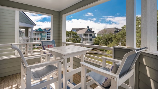 A covered outdoor patio with white furniture, a view of neighboring houses, and a wooden deck.