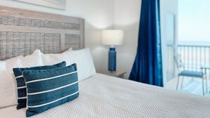 Ein ordentlich bezogenes Bett mit weiß-blauen Kissen, einer weißen, strukturierten Bettdecke, einem Holzkopfteil, einer Nachttischlampe und Blick auf einen Balkon mit einem Stuhl.
