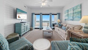 Ein Wohnzimmer mit Fernseher, Deckenventilator, Sofa, Couchtisch und Blick auf den Strand durch große Fenster.