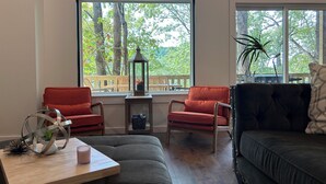 Salón con dos sillas rojas, un sofá gris, una mesa de madera con una vela y un farol. Amplias ventanas ofrecen vistas a árboles y una terraza exterior.
