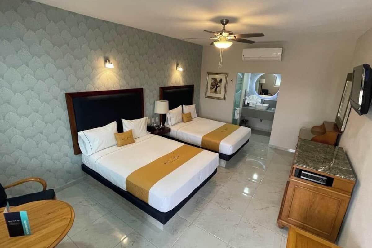 Une chambre d’hôtel avec deux lits, un ventilateur de plafond, une télévision, un bureau et une salle de bain visible au fond.