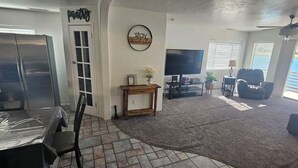Una sala de estar con televisor, un comedor con mesa y sillas, una consola de madera con un florero, y un balcón con vistas.
