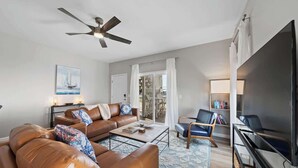 A living room with a brown leather sofa, a coffee table, a television, and a ceiling fan. - Carolina Breeze by Carolina Getaway - Spacious 3BR Condo w/ Ocean & Sound Views (Carolina Beach)