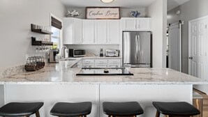 A modern kitchen with white cabinets, a marble countertop, a central island with stools, a refrigerator, and a microwave. - Carolina Breeze by Carolina Getaway - Spacious 3BR Condo w/ Ocean & Sound Views (Carolina Beach)