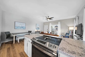 A modern kitchen with a built-in stove, a dining area with a bench, and a living space with a sofa and a TV. - Carolina Breeze by Carolina Getaway - Spacious 3BR Condo w/ Ocean & Sound Views (Carolina Beach)
