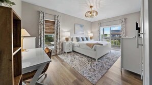 A bedroom with a bed, bedside tables, a desk, and a view of the outside. - Carolina Breeze by Carolina Getaway - Spacious 3BR Condo w/ Ocean & Sound Views (Carolina Beach)