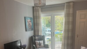 Un salon avec un canapé, un fauteuil et un téléviseur posé sur un meuble en bois. Il y a un tableau au mur et une fenêtre avec des rideaux.
