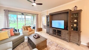 A living room with a large flat-screen TV, a ceiling fan, and a view of a green landscape through large windows.