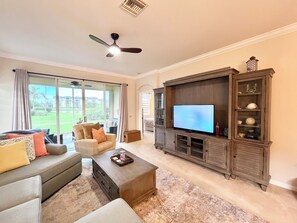 Een woonkamer met een grote flatscreen-tv, een plafondventilator en uitzicht op een groen landschap door grote ramen.