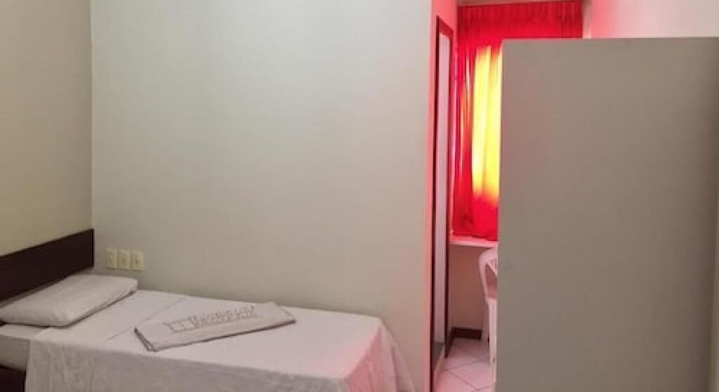 A small, single-bed room with a white bedspread and a pillow. There is a small table with a piece of paper on it. The room has a plain white wall and a door with a red curtain.