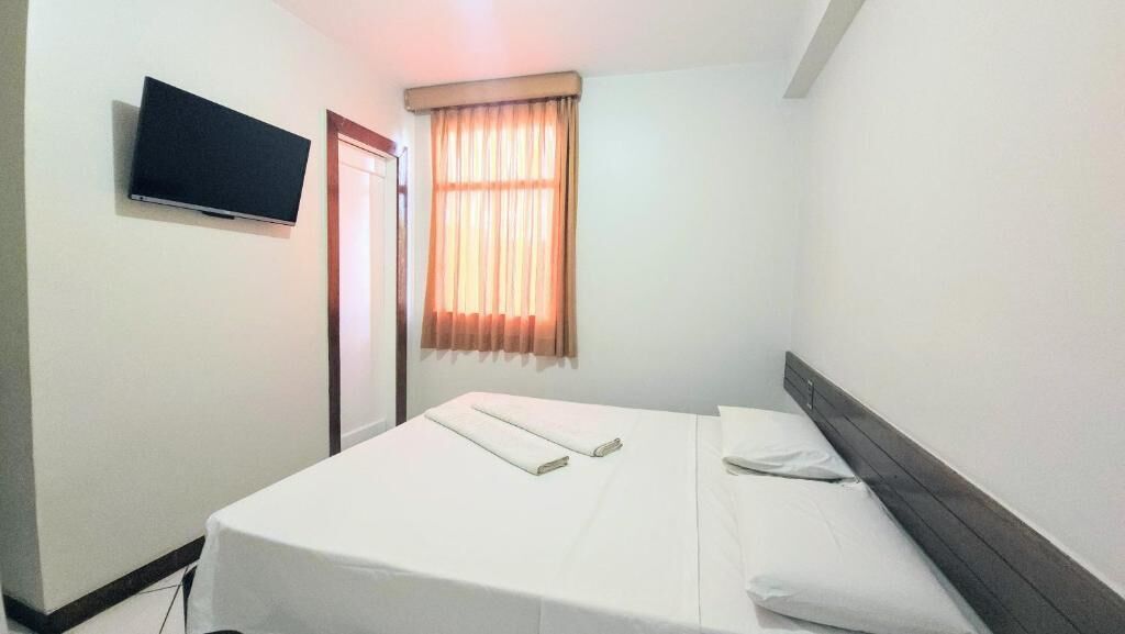 A hotel room with a bed, a television mounted on the wall, a window with curtains, and a door.