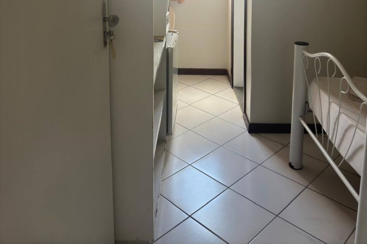 A narrow hallway with a bed on the right, a window with curtains, and a door on the left.