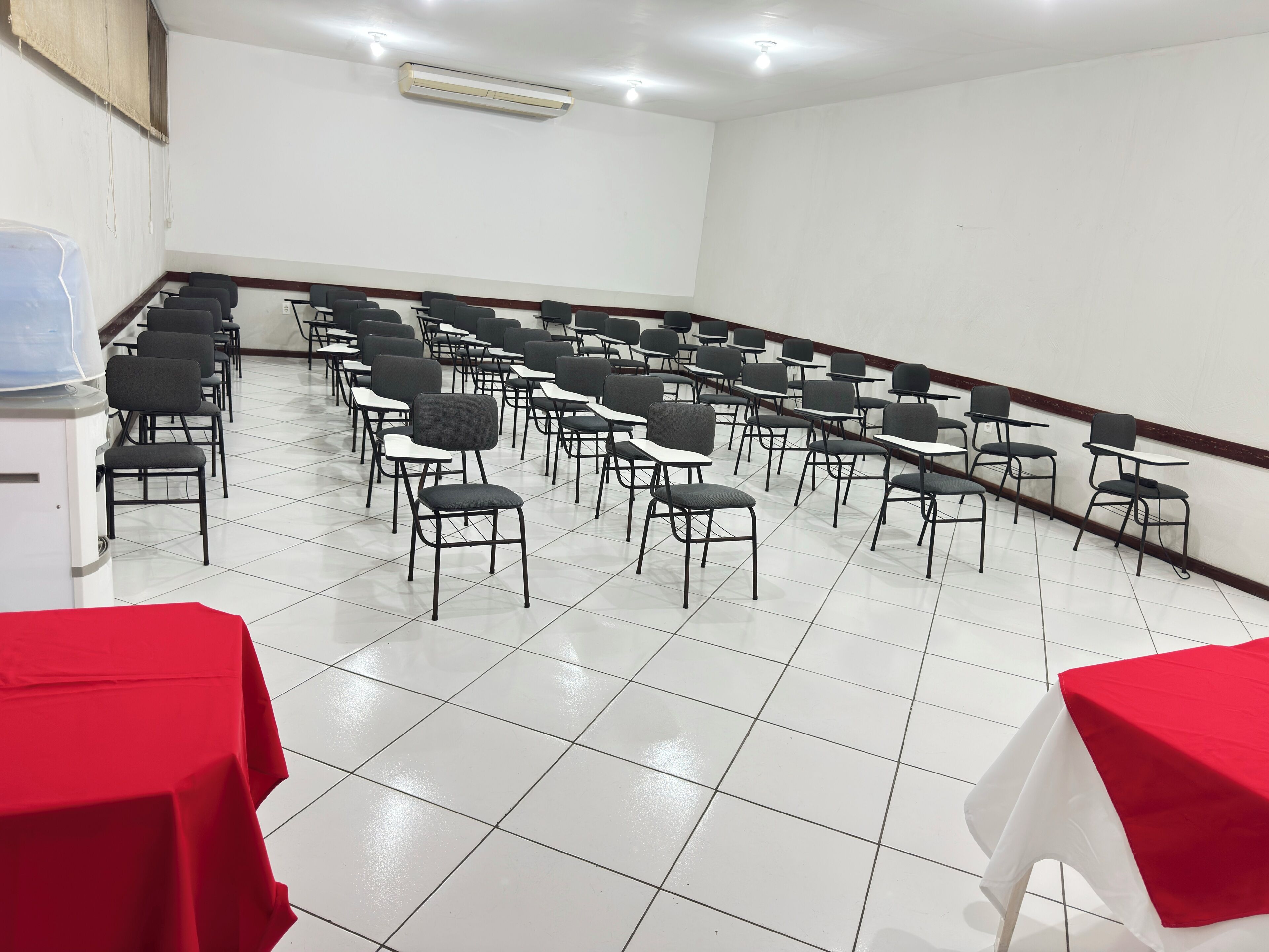 Un salón de conferencias con filas de sillas y mesas, paredes blancas y piso de baldosas.