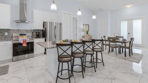 A modern kitchen with a central island, bar stools, a dining table, and a large window.