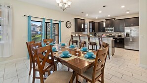 Una cocina moderna con armarios oscuros, electrodomésticos de acero inoxidable y un comedor con sillas de madera y vajilla azul.