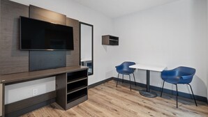 Een moderne kamer met een flatscreen-tv aan een houten wand, een klein plankje en een tafel met twee blauwe stoelen.