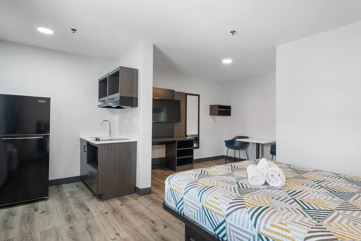 A modern hotel room with a kitchenette, a bed with a patterned bedspread, and a desk with a chair.