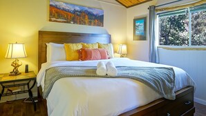 Una camera da letto ben illuminata con soffitto in legno, un letto grande con molti cuscini, comodini con lampade e un quadro che raffigura un paesaggio montuoso.