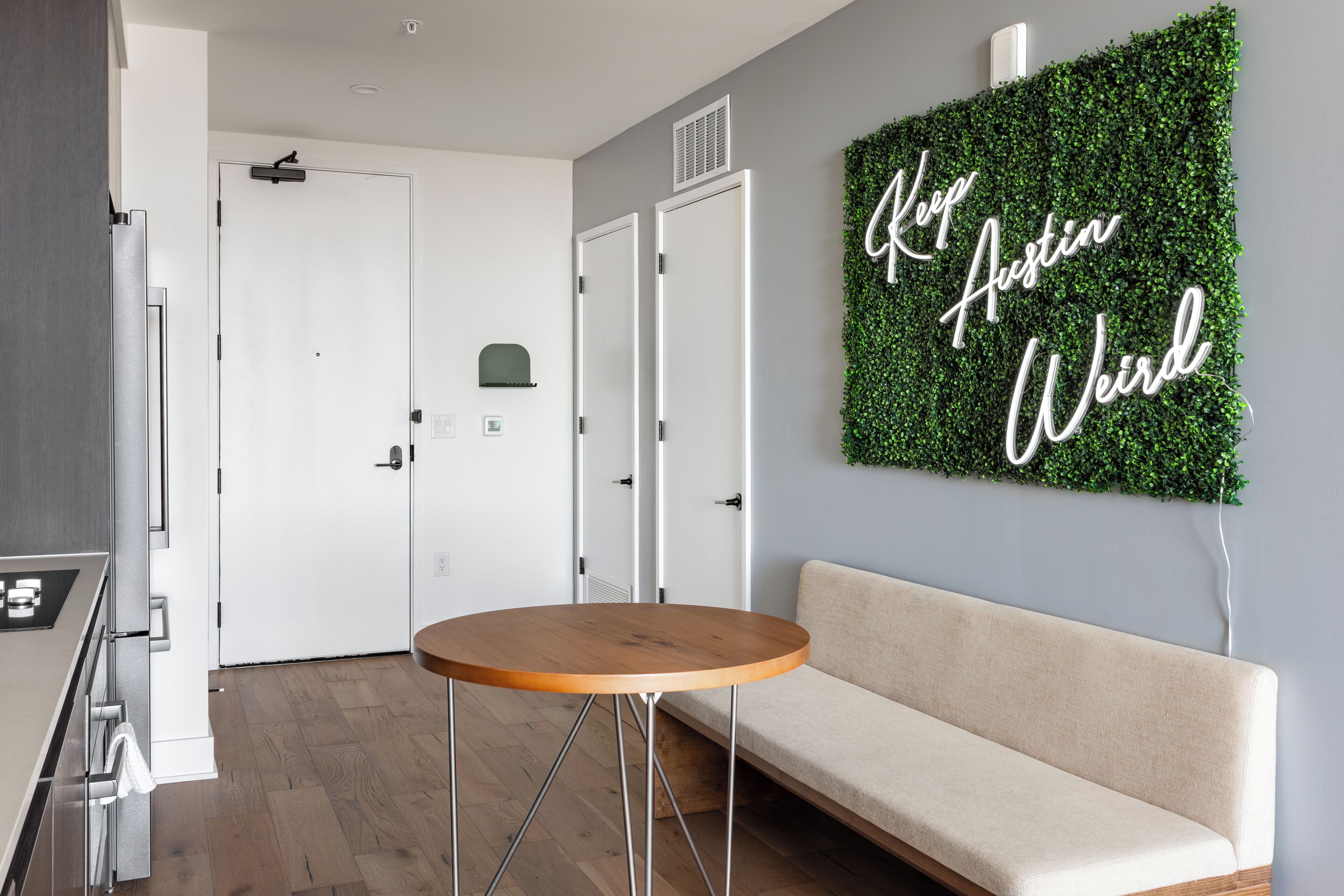 A modern interior with a round wooden table, a beige sofa, and a wall-mounted green foliage sign with the text "Keep Austin Weird."