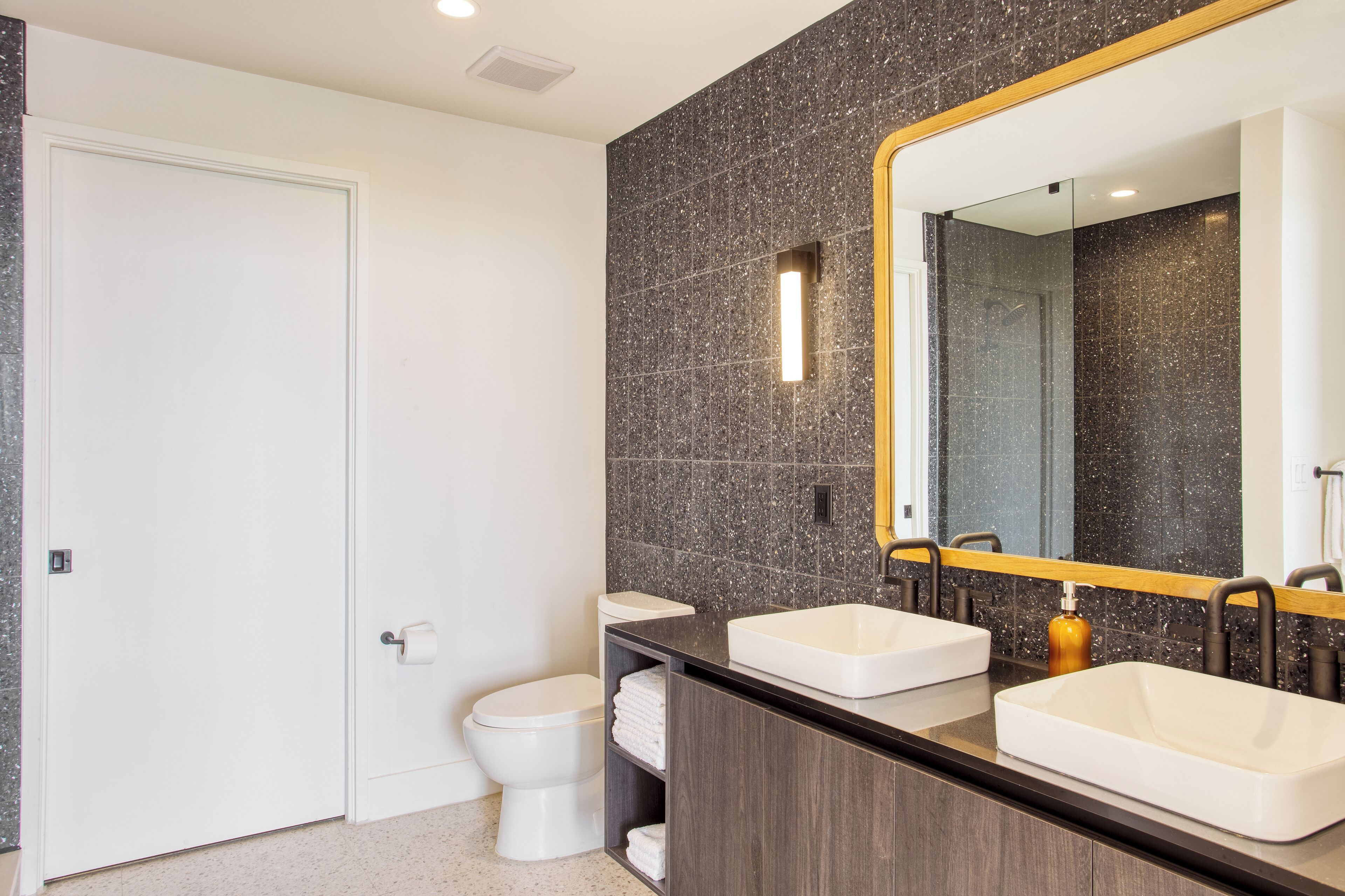 Une salle de bain moderne avec des toilettes blanches, deux lavabos, un grand miroir et des carreaux foncés à motifs.