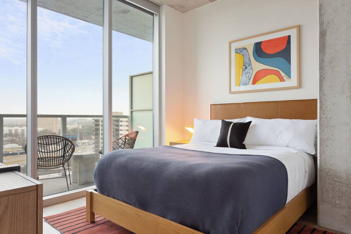 A modern hotel room with a large bed, a view of the city through floor-to-ceiling windows, a balcony with a chair, and a colorful abstract painting on the wall.