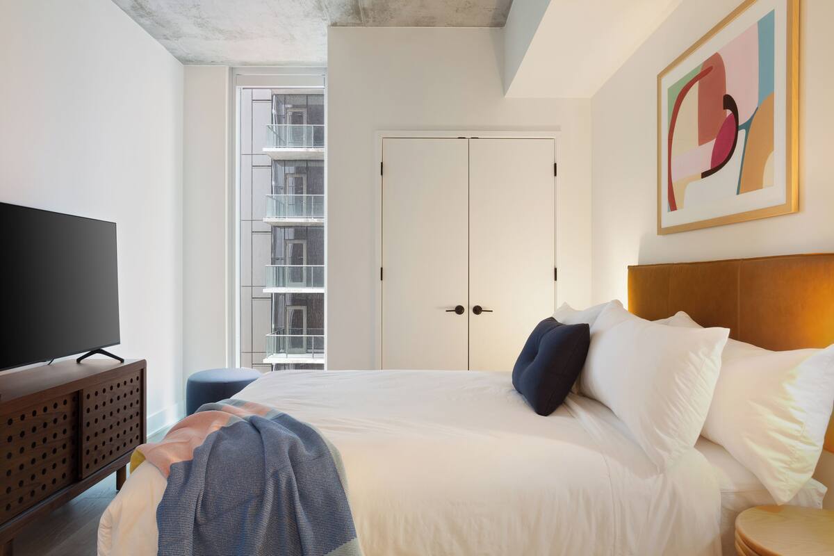 A bedroom with a bed, a television on a wooden cabinet, a window with a view of buildings, and a door.
