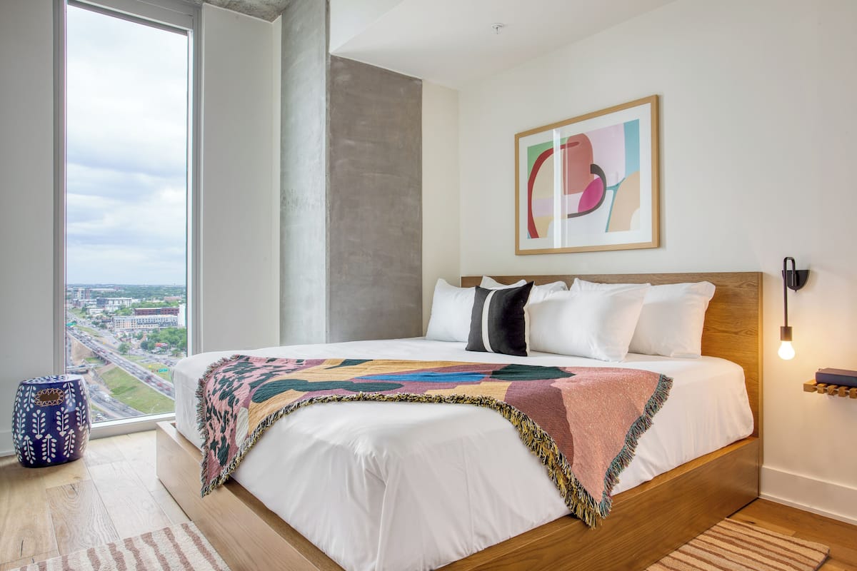 A modern bedroom with a large bed, a view of the city, and a colorful abstract painting on the wall.