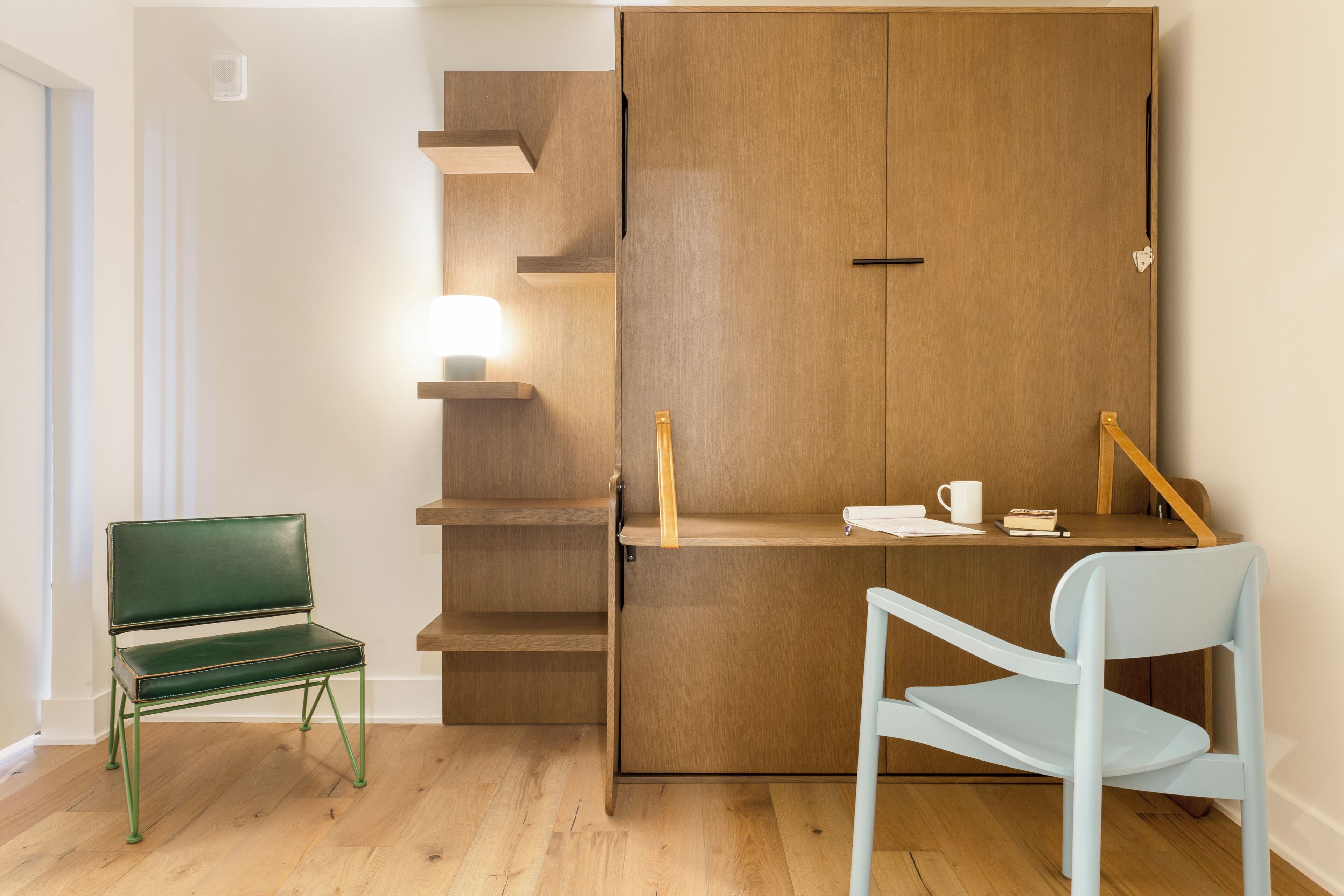 A room with a wooden wardrobe, a desk with a chair, a lamp, and a bookshelf.