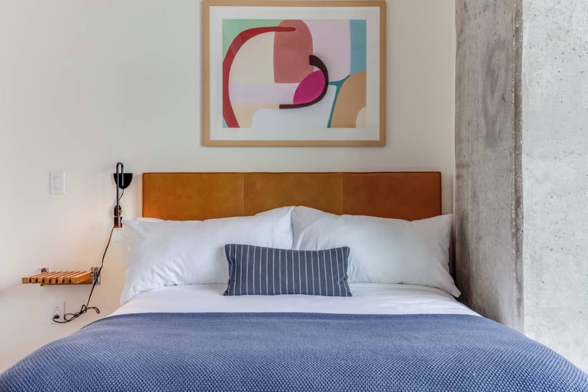 A neatly made bed with a blue blanket, white pillows, and a striped pillow. A framed abstract artwork hangs above the bed.