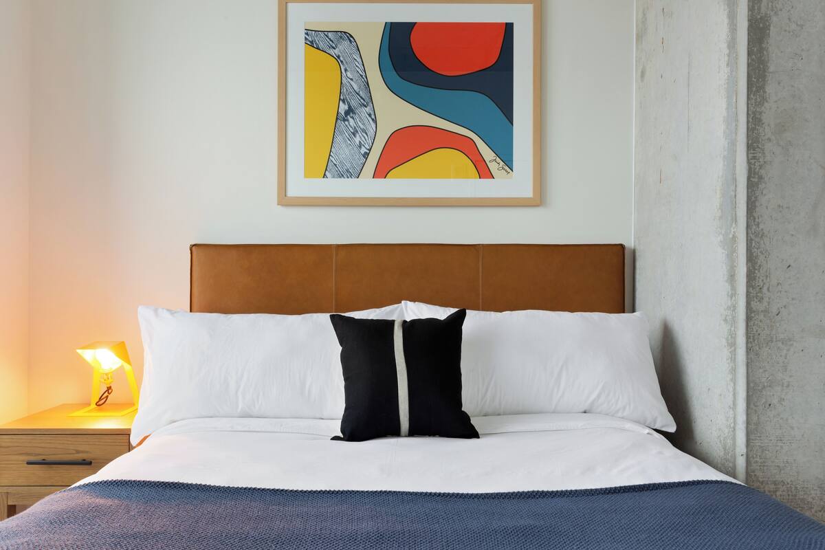 A neatly made bed with white and black pillows, a blue blanket, and a framed abstract painting above it.
