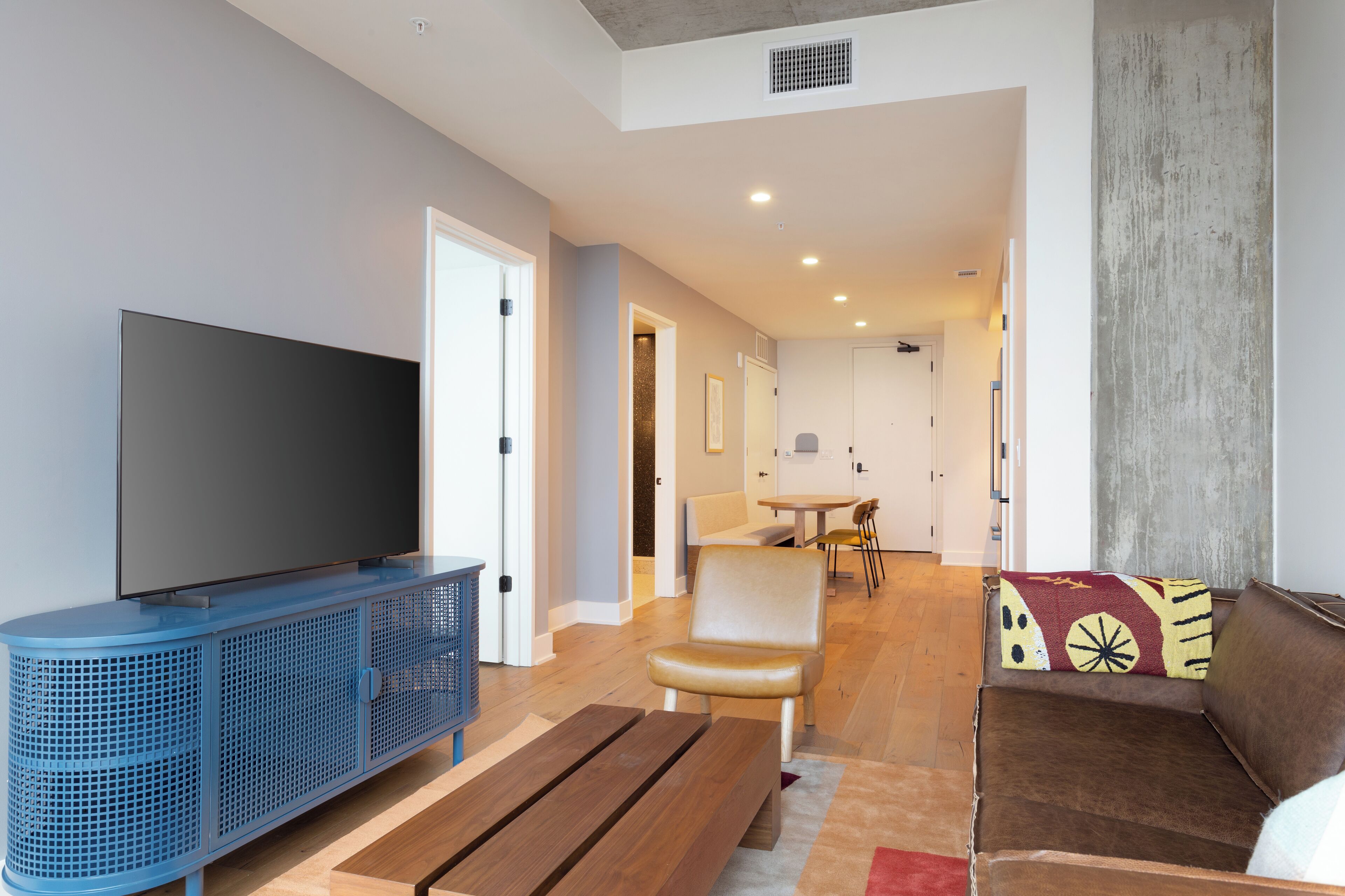 A modern living room with a flat-screen TV, a blue entertainment unit, a wooden coffee table, a brown sofa with a patterned throw, and a dining area in the background.